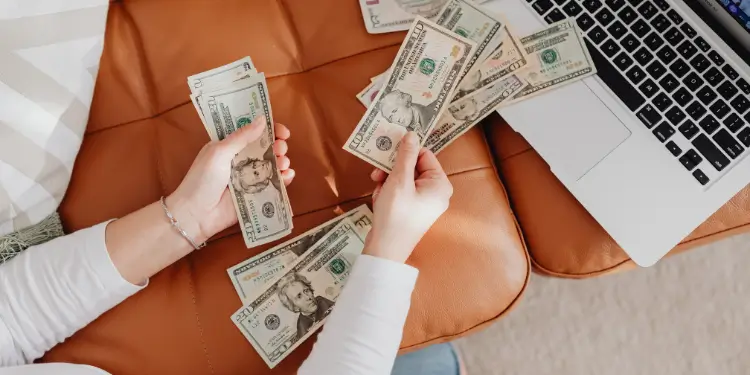 Top-down shot showing hands handling multiple dollar bills next to an open laptop keyboard.