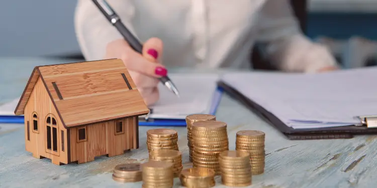 Wooden house model with coins and paperwork showing housing expenses encroaching on budget needs.