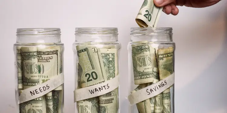 Photo of three dollar-filled jars labeled Needs, Wants, Savings with a hand adding a bill.
