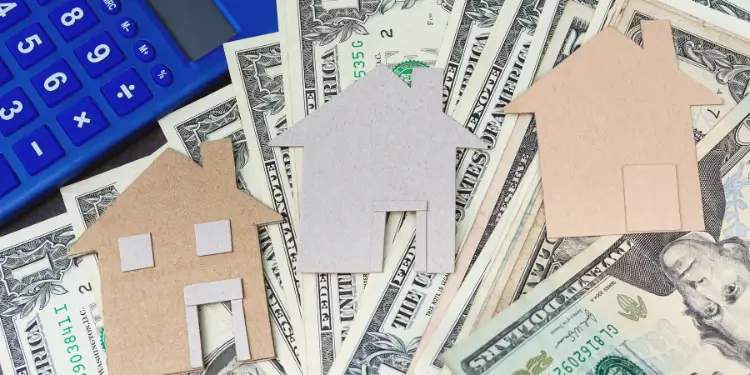 Cardboard houses placed on cash with calculator to represent lowering housing costs via roommates.
