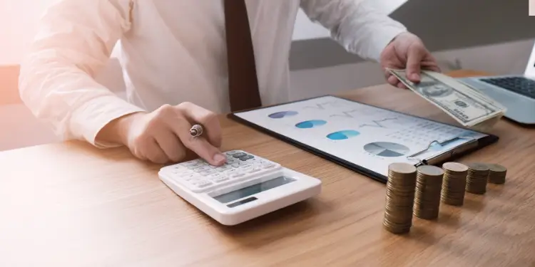 Hands holding a calculator with cash, coin stacks, and financial charts to review take-home pay accuracy.