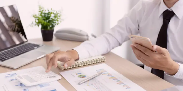 Person in shirt and tie at desk reviewing charts with calculator and smartphone to curb overspending.