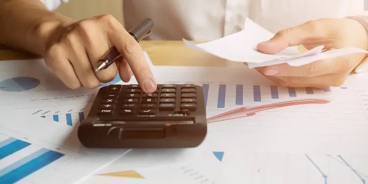 Hands using a calculator with financial charts to allocate monthly sinking funds for annual expenses.