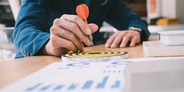 Hand placing a dart on a bullseye over charts to symbolize income variability affecting budgets.